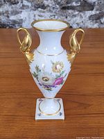 Full front view of the white porcelain vase displaying floral painting and gilded swan handles.
