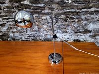 Chrome ball adjustable table lamp standing on wooden table with stone wall background, showing profile view and chrome finish.