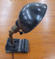 Black metal industrial clamp lamp with rounded shade, adjustable arm, and clip base shown from side angle on wooden surface.