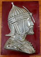 Front view of a cast metal knight plaque mounted on a red metal backplate, showing detailed armor and helmet relief.