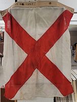 Full view of military ship flag with red diagonal cross, showing canvas material, grommets, and stamped marking "FLAG V SIZE 6 1957" on edge.