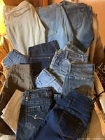 12 pairs of women's jeans and pants folded and laid out showing various denim washes and styles, including light, medium, and dark blue, black and cream tones, taken to show full lot contents.