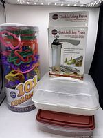 Boxed unused cookie/icing press next to large container of colorful cookie cutters and two stacked baking containers, one clear with white lid and one with red lid.