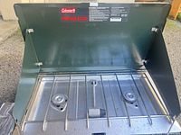 Open view of green Coleman propane stove showing 2 burners with protective metal grate and foldable windshield sides.