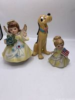Photo showing the set of three figurines: Disney Pluto standing in the center, flanked by two July birthday angel figurines, one holding a flower and the other an American flag.