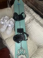 Top view of one snowboard in light blue with attached black bindings and pair of white snowboard boots