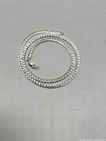 Full view of the silver chain necklace laid coiled on a surface, showing the flat interlocking link design and lobster claw clasp.