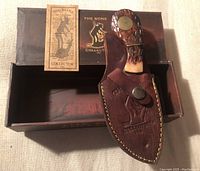 Cutting tool in leather sheath with original box and paperwork on beige fabric background.