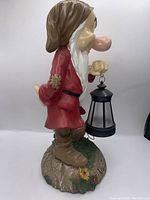 Side profile showing Grumpy in red robe holding black lantern. Detail on base with flowers.