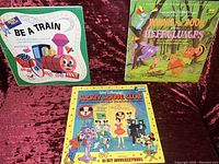 Three vintage vinyl LP albums: 'Be A Train,' 'Winnie the Pooh and the Heffalumps,' and 'Mickey Mouse Club' on red velvet background.