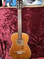 Front view of vintage acoustic guitar showing wear and natural finish, resting against a burgundy fabric background.