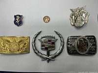 Top view of collection showing six items: 2 pins, 2 buckles, Cadillac emblem, and penny for scale.