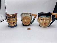 Front angle of three vintage Toby style character mugs and creamers, showcasing their detailed hand-painted faces and shapes, with a penny for scale.