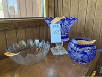 Three glass items displayed on wood surface including cobalt blue cut glass round bowl, cobalt blue cut glass vase on a clear pedestal, and an opaque flower petal shaped bowl.