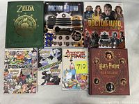 Overview photo showing all seven books: The Legend of Zelda Hyrule Historia, two Doctor Who books, two Pokemon Black & White Pokedex volumes, Adventure Time Volume 3 graphic novel, and Harry Potter Film Wizardry book.