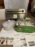 Photo showing FoodSaver vacuum sealer, marinating dish with lid, boxed sealing rolls, and printed manuals displayed on floor.