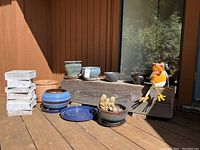 Wide shot of all garden pots, saucers, decorative stack of white books, metal frog figure, and cactus plant in pot on wood deck. Items show outdoor wear and slight dirt.