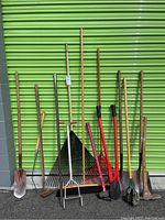 Wide view of various large garden and hand tools standing upright against green metal backdrop