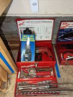Overview of the open red Milwaukee metal case with hammer drill, drill bit sharpener box, and various drill bits.