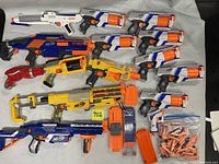 Image showing entire collection of 14 Nerf blasters laid out on table with stocks, magazines, and darts. Includes multiple Strongarm blasters, yellow Maveric Rev-6, blue Retaliator and Recon CS-6, white blasters, and orange dart clips and darts.