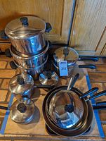 Front view of the set of pots and pans arranged on a brick floor, showing multiple stainless steel pots and sauce pans with matching lids and black handles.