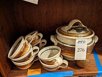 Front view of set showing the large pottery soup tureen with lid, stacked bowls, and copper ladle alongside in wooden shelving.