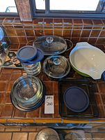 Full view of the lot showing clear Pyrex glass bowls, two clear glass casserole dishes with lids, green and white mixing bowl, black plastic-lidded dishes, and other glassware.