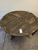 A round wooden outdoor table top with slatted design and weathered surface, showing a central hole for umbrella.
