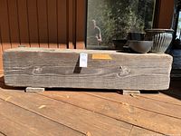 Front and side view of a long rustic solid wood bench placed outdoors on a wooden deck, showing the weathered wood surface and wood feet supports.