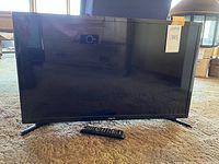 Front view of the Samsung 32-inch TV powered off with remote control placed in front on carpet.
