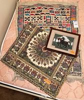 Three textiles laid on a bed: framed needlepoint horses, vintage woven textile with geometric patterns, ornate embroidered textile with gold thread medallion design.