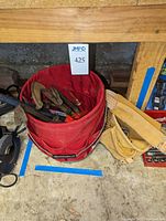 Photo of red fabric bucket holding assorted clamps and tools by floor and a leather tool belt to the right