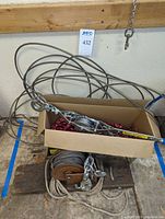 Photo showing hand winch with wire cable lying in a cardboard box with red and black rope underneath, coiled wire cable visible around it.