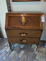 Front view of oak wood drop-front secretary desk with floral carved decoration and two drawers with brass pulls.
