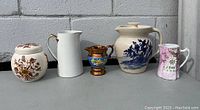 Photo showing five antique porcelain items placed side by side: ginger jar, white creamer, copper-tone pot, Staffordshire jug, and pink floral creamer.