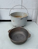 Shows two cast iron pots, one gray and one white, both with wire handles for hanging, placed on a gray surface in front of a white brick wall.