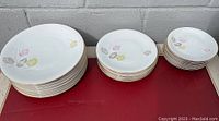 Stacks of large, medium, and small bowls and plates with pastel leaf pattern and gold rim on a red surface outdoors.