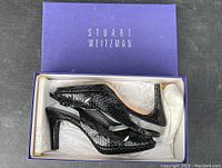 Pair of black snakeskin patterned slingback shoes inside an open Stuart Weitzman brand shoe box, showing the style and heel shape.