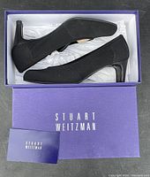 Pair of black knit Stuart Weitzman pumps displayed inside original purple box with white tissue paper, showing side profile of shoes and sole.