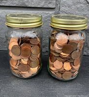Two glass fruit sealer jars filled with vintage Canadian pennies shown side by side on a dark surface against a rough gray wall.