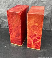 Pair of rectangular dyed marble bookends photographed against a gray stone wall and dark surface, showing their polished, glossy red and orange veining and overall shape.