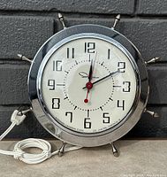 Front view of the vintage Ingraham chrome wall clock showing dial and design with ship wheel style spokes and power cord.