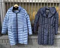Both winter coats hanging side by side on a metal railing showing their full fronts. Left coat is metallic blue, right coat is black.