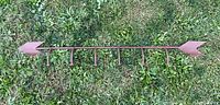Full length view of the wrought iron arrow shaped wall hanging with six coathooks placed on grass, showing its length and arrow shape