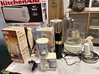 Photo showing all kitchen appliances together on table including boxed Braun Multiquick hand blender, KitchenAid toaster box, Cuisinart food processor, Braun coffee machine, peppermill, Osterizer blender, Krups mixer.