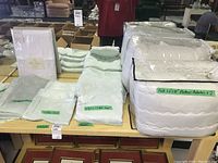 Photo showing two full size mattress protectors in packaging, queen flat and fitted sheets, pillow cases, all light green or white, labeled on a table.