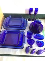 Full lot view showing sets of cobalt blue glassware on a white surface against a green and white background, including baking dishes, stemware, bowls, plates, and small decorative pieces.