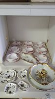 Multiple Victorian Violets patterned tea cups and saucers arranged on shelf along with small decorative plates and a footed serving bowl with gold floral decoration and utensils.