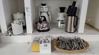 Overview of white and stainless steel coffee makers, electric kettle, juicer, boxed cheese graters and basket of flatware on shelving