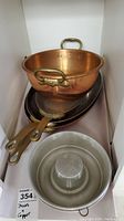 Stacked cookware including two copper Bundt pans with tin interiors and copper exterior, multiple brass handled skillets beneath, and metal bowls in the foreground.
